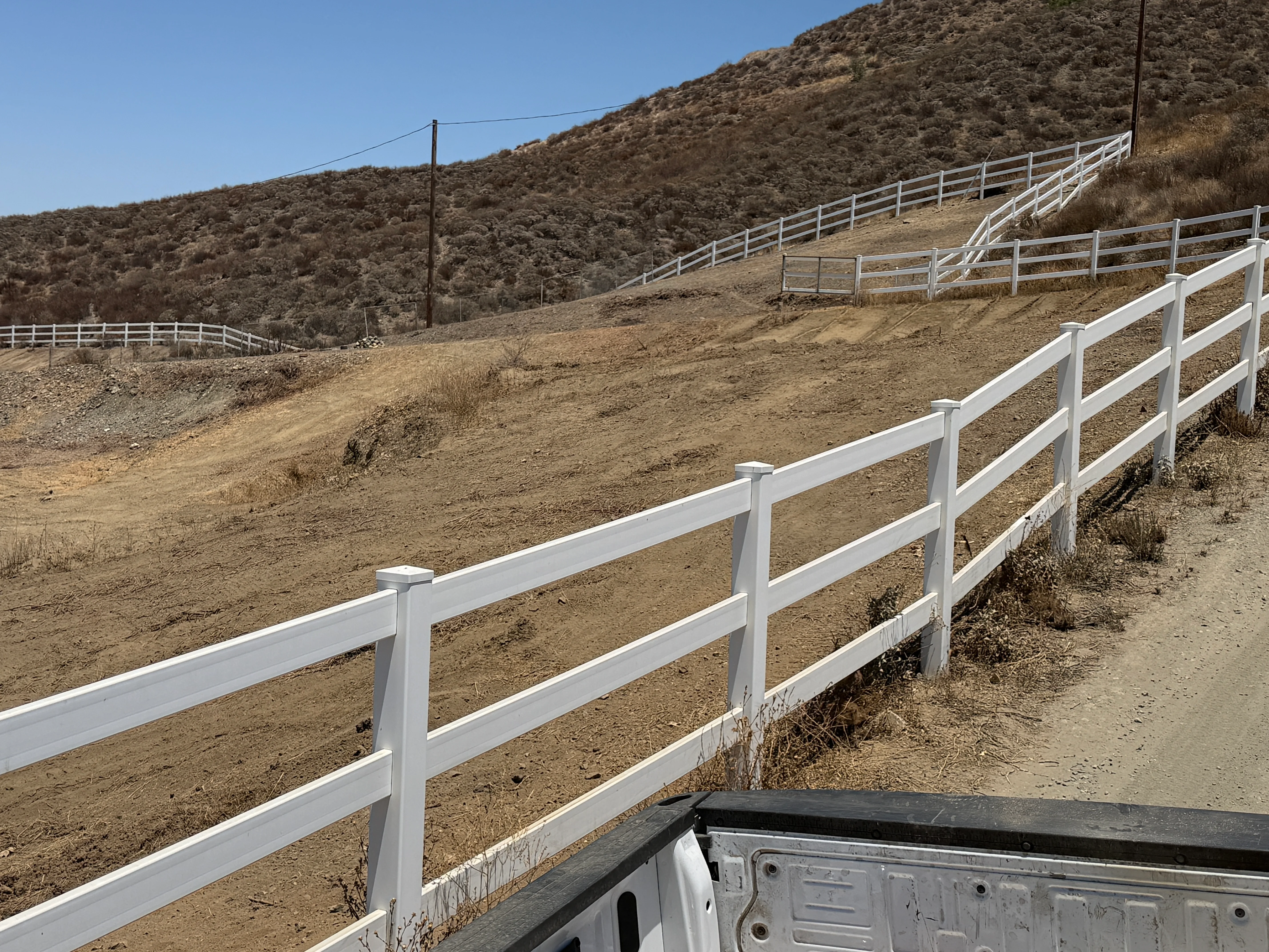 White ranch fencing on cleared hillside property with sparse vegetation and dirt terrain
