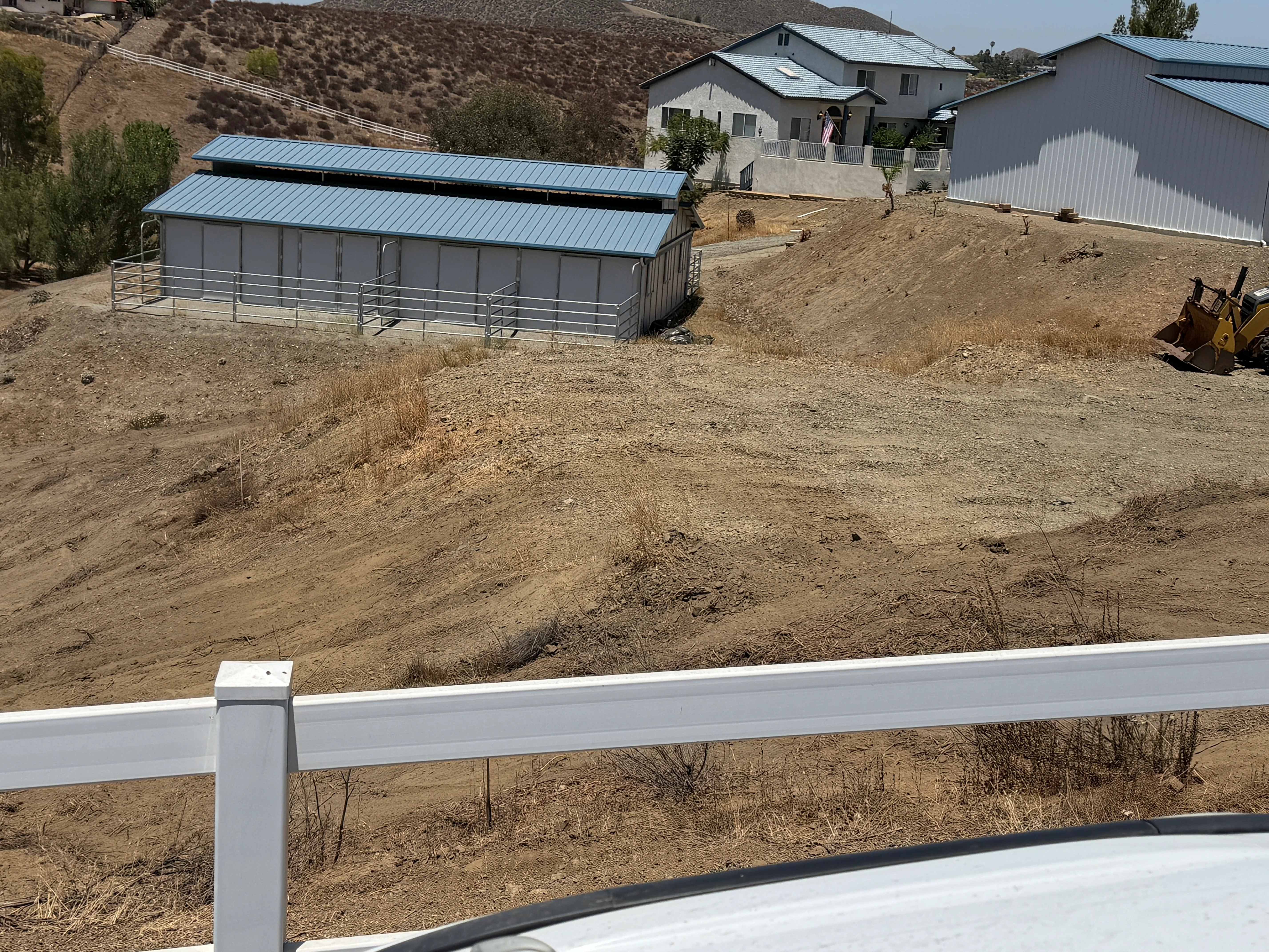 Cleared rural property with graded dirt, white vinyl fencing, and metal-roofed structures on hillside