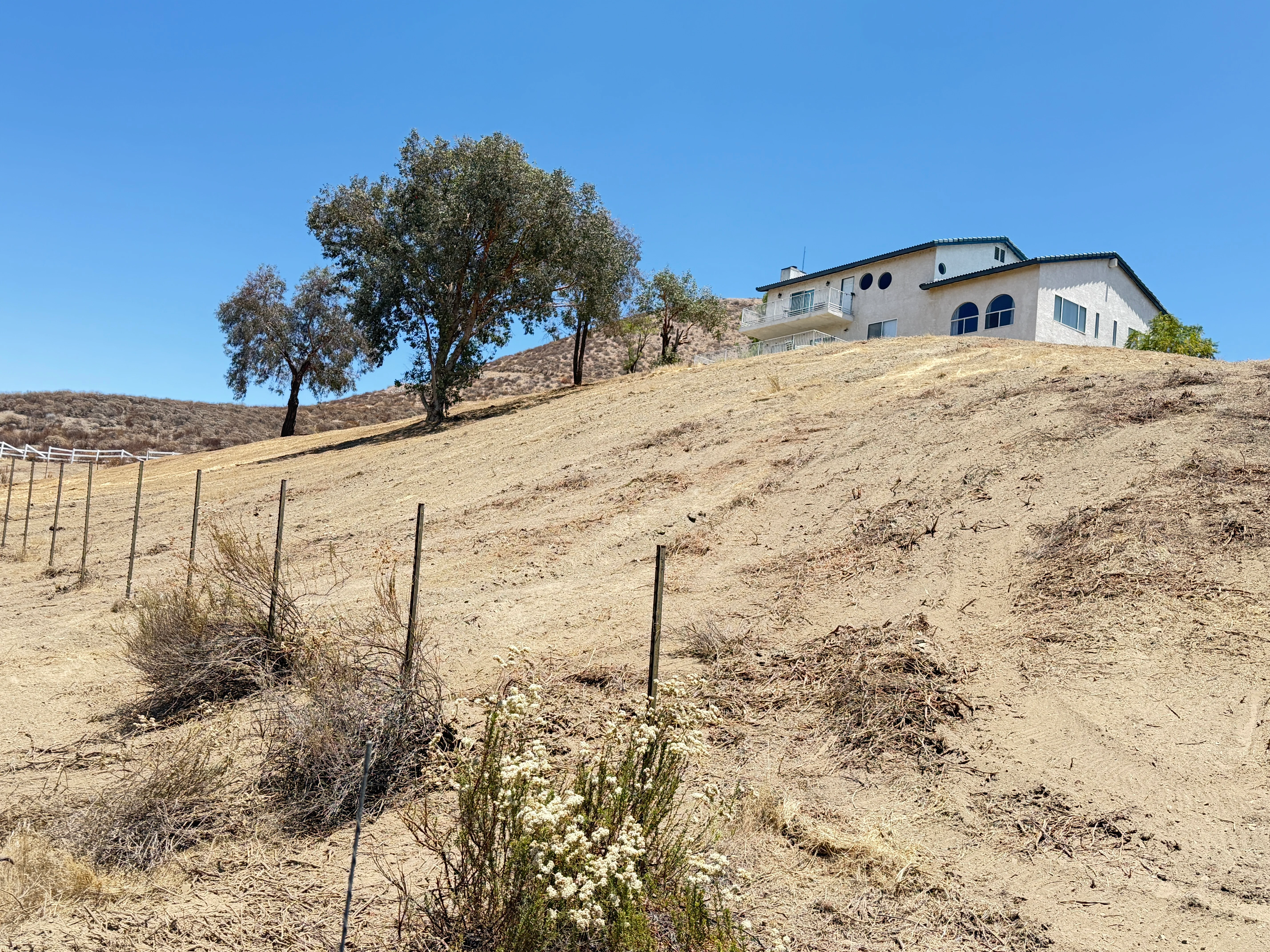 Cleared hillside with erosion control fencing, native plantings, and white Mediterranean-style home on ridge