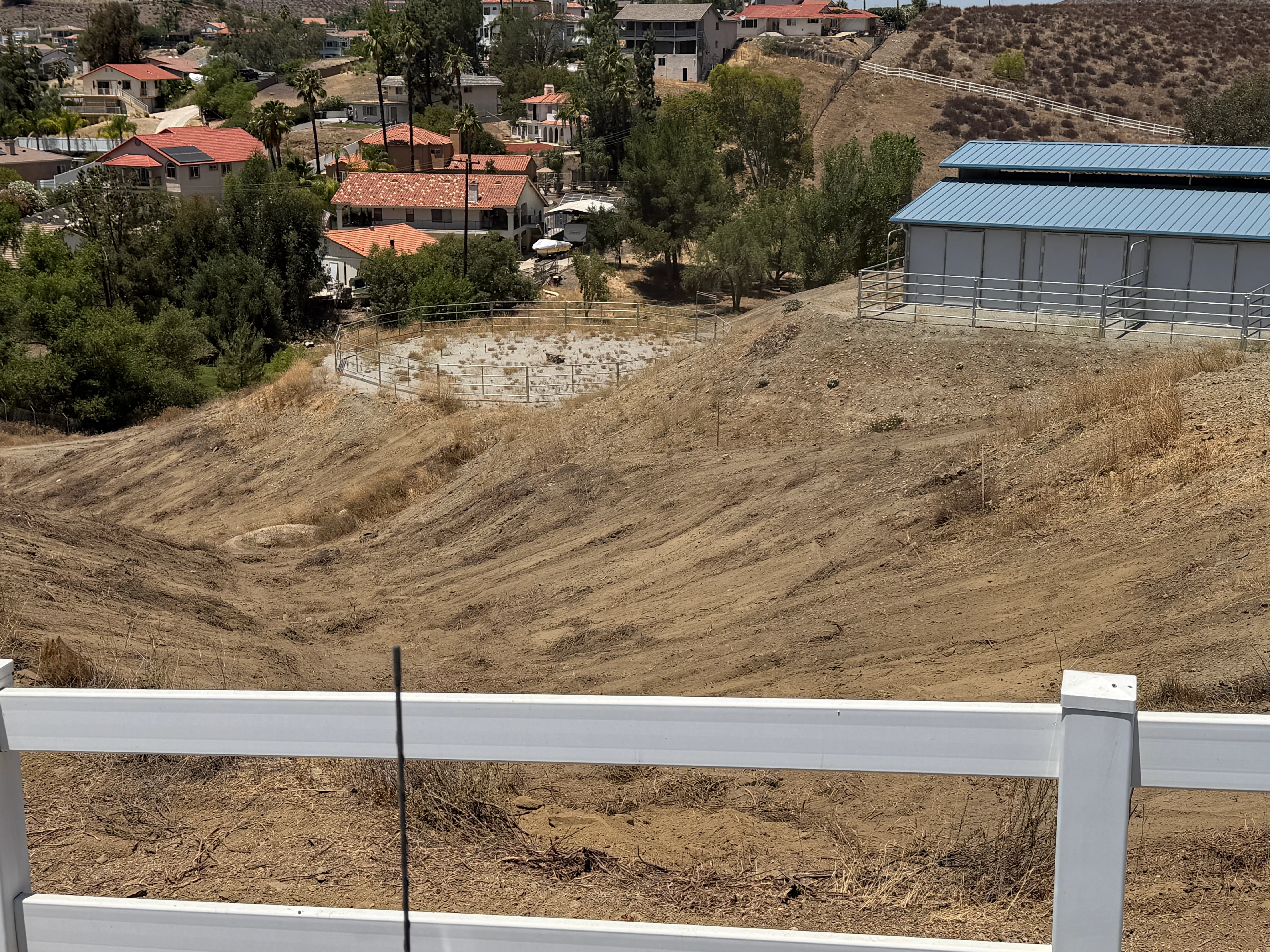 Cleared hillside property with white ranch fencing and metal storage building in background